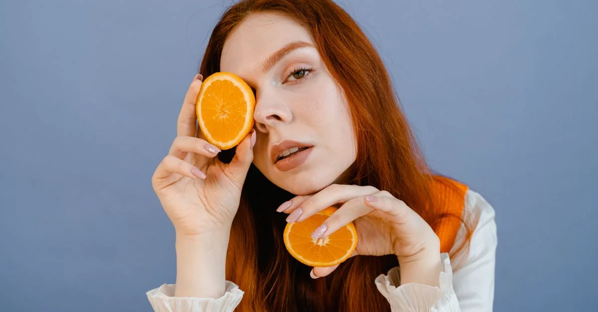 Portrait vibrant d'une jeune femme aux cheveux roux tenant des tranches d'orange sur un fond bleu.