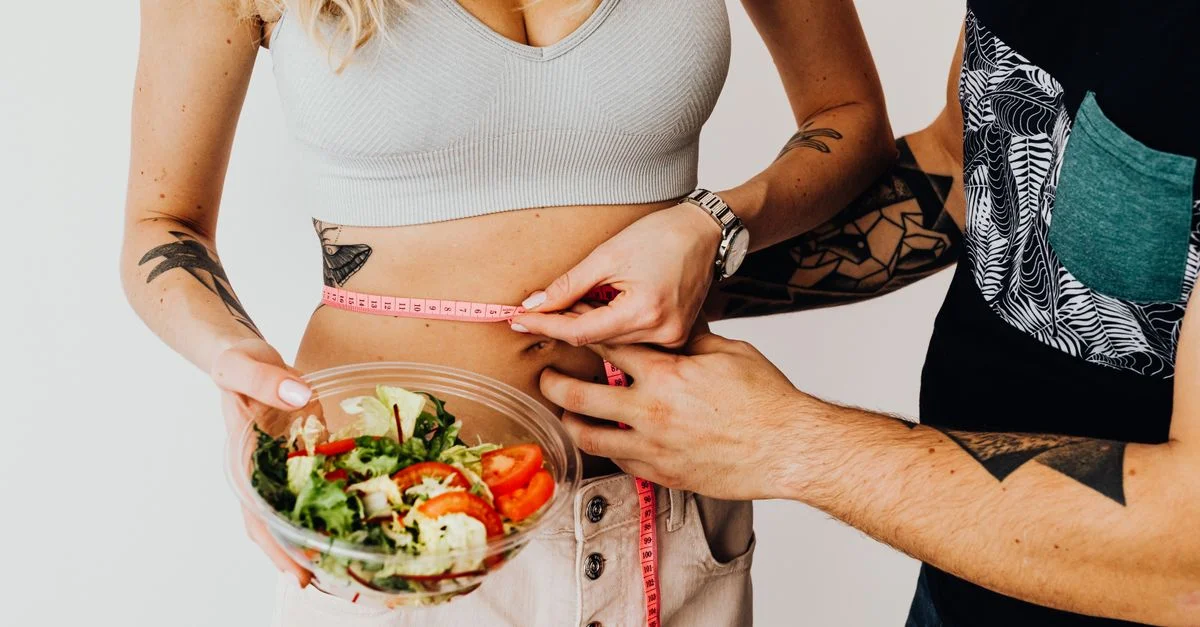 Un couple mesurant la taille avec un ruban et tenant un bol de salade fraîche, axé sur la forme physique et la perte de poids.