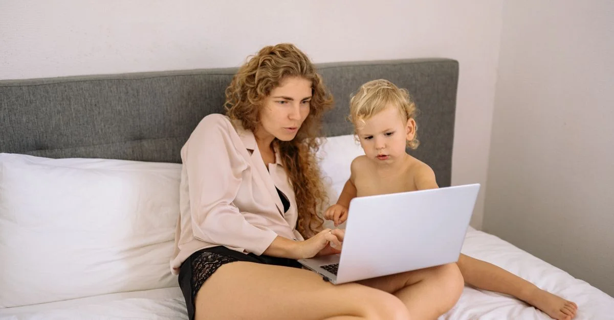 Una madre y su niño pequeño disfrutan de tiempo de calidad usando un laptop juntos mientras están sentados en una cama.