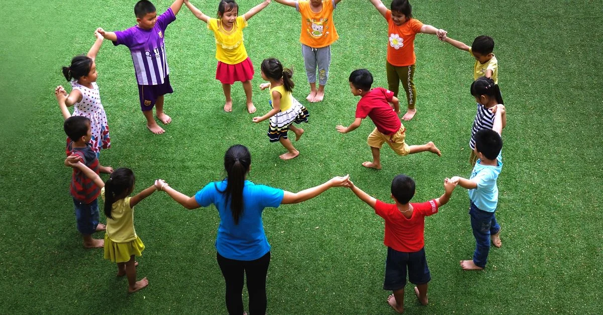 Niños tomados de la mano en círculo sobre el césped verde, participando en un juego divertido al aire libre.