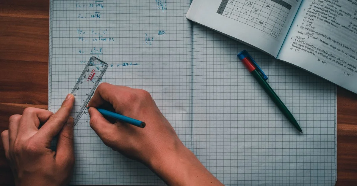 Una persona escribe ecuaciones matemáticas en un cuaderno usando una regla y un lápiz, centrada en resolver problemas.