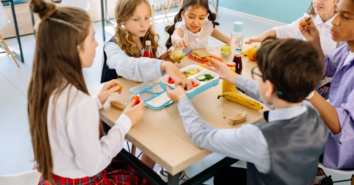 Grupo de niños diversos almorzando en una cantina escolar, compartiendo y disfrutando de bocadillos saludables.