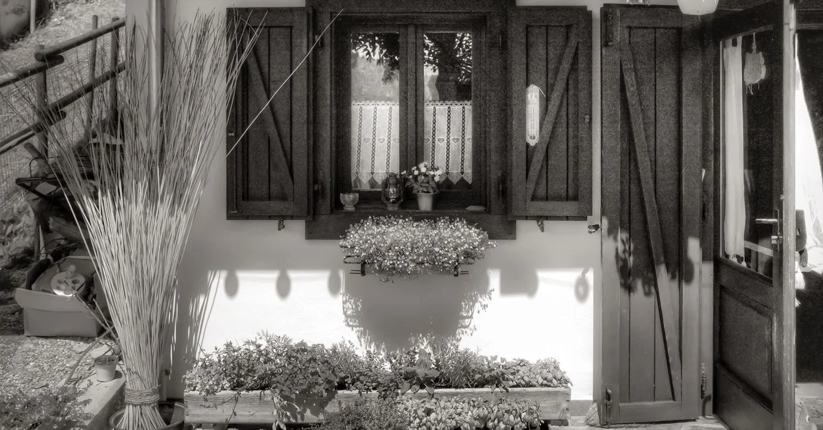 Photo en noir et blanc d'une cour italienne avec volets en bois, fleurs et lumière du soleil.