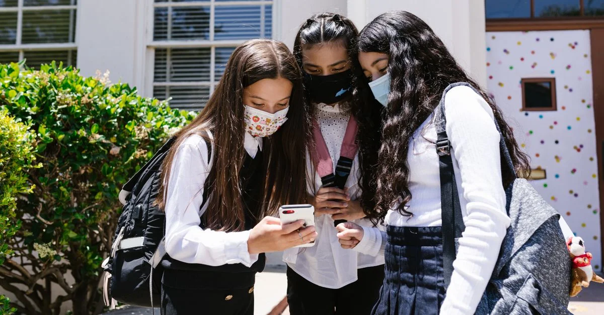Tres estudiantes en uniforme usando mascarillas y utilizando un teléfono móvil fuera de un edificio escolar.