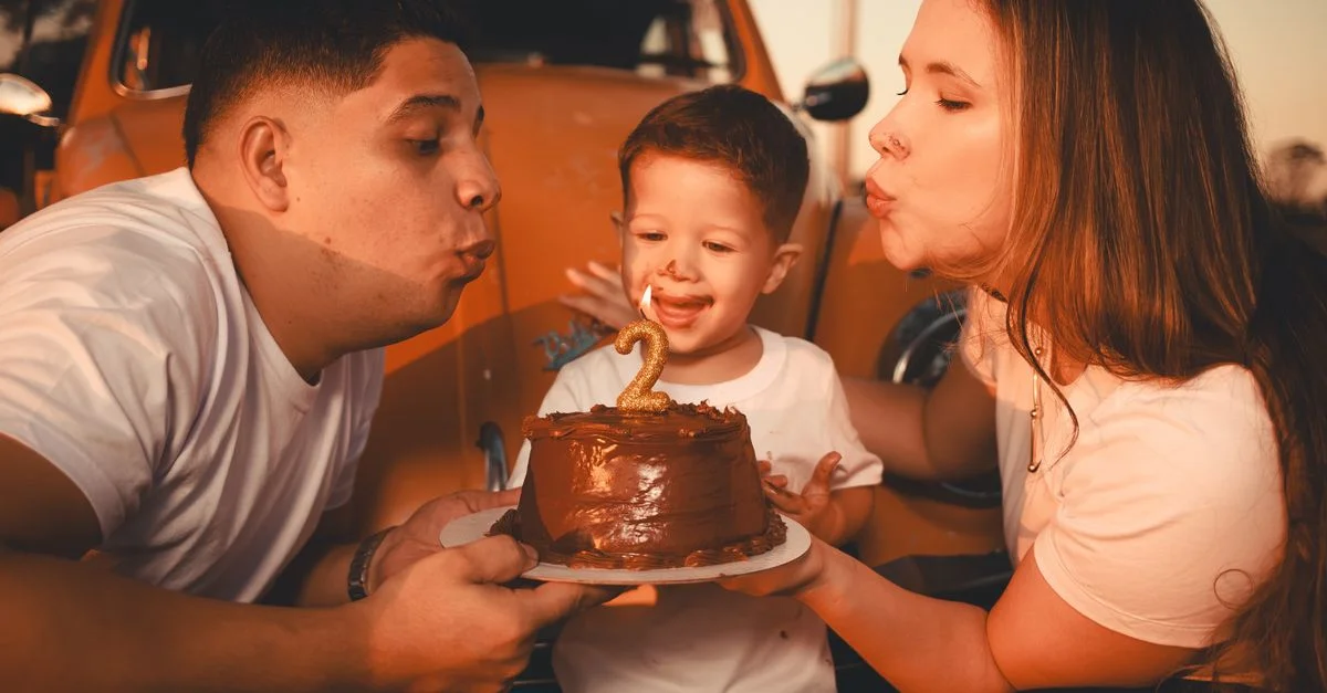 Familia de tres personas celebrando el segundo cumpleaños de un niño con un pastel al aire libre.