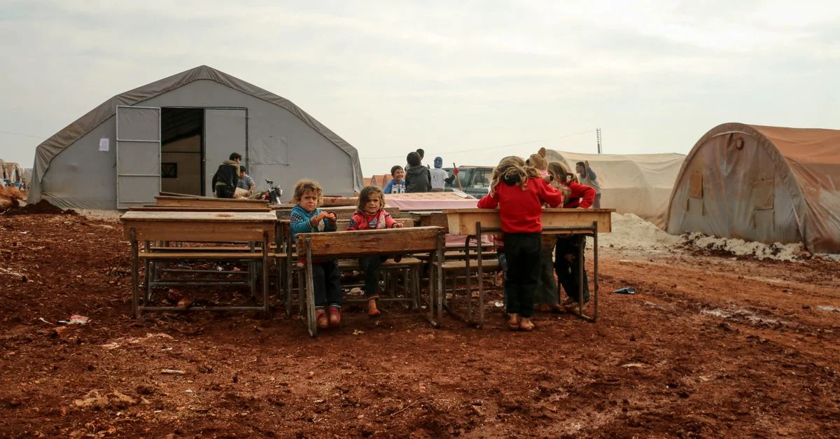 Des enfants se rassemblent autour de bureaux dans un camp de réfugiés à Idlib, en Syrie, soulignant la résilience et la communauté.