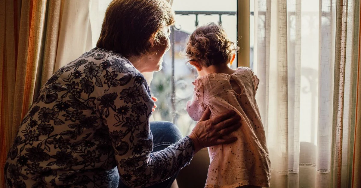 Un momento de amor entre abuela y nieto junto a una ventana llena de luz solar.
