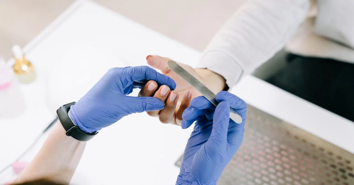 Acercamiento de una técnica de uñas limando las uñas de una cliente con guantes azules en un entorno profesional.