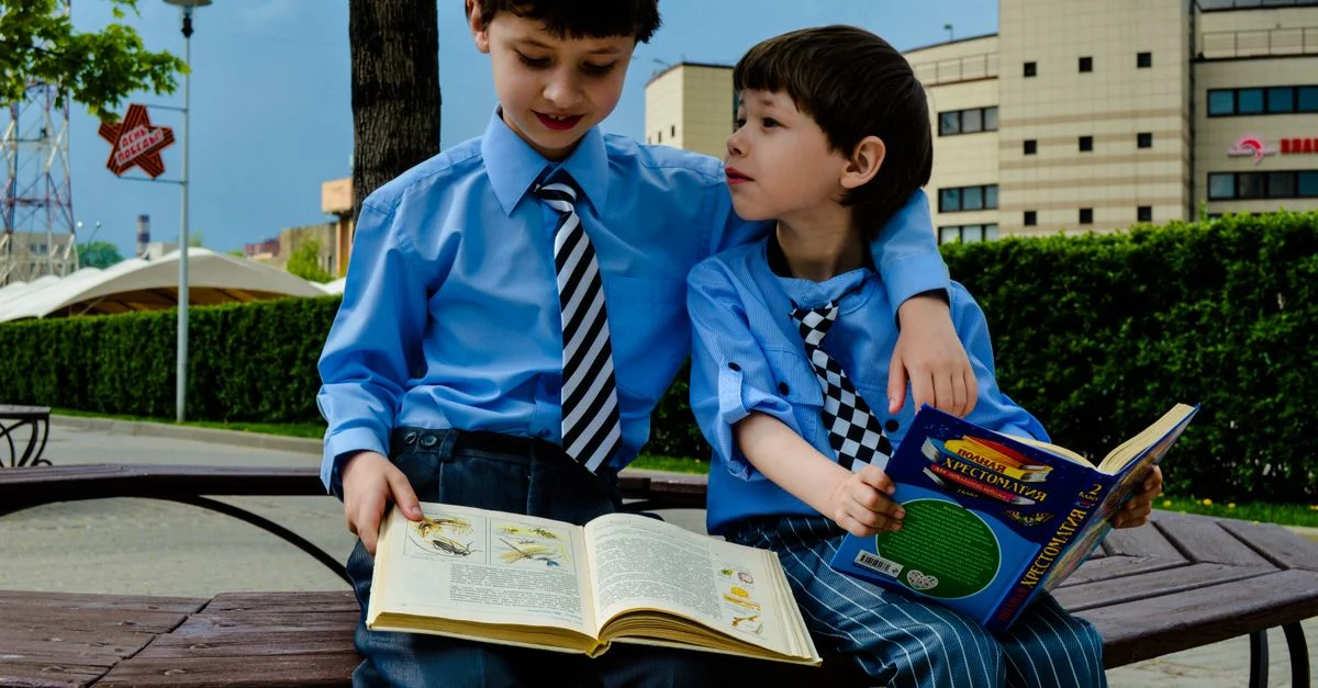 Dos niños pequeños con corbata se sientan en un banco leyendo libros en un parque urbano soleado.