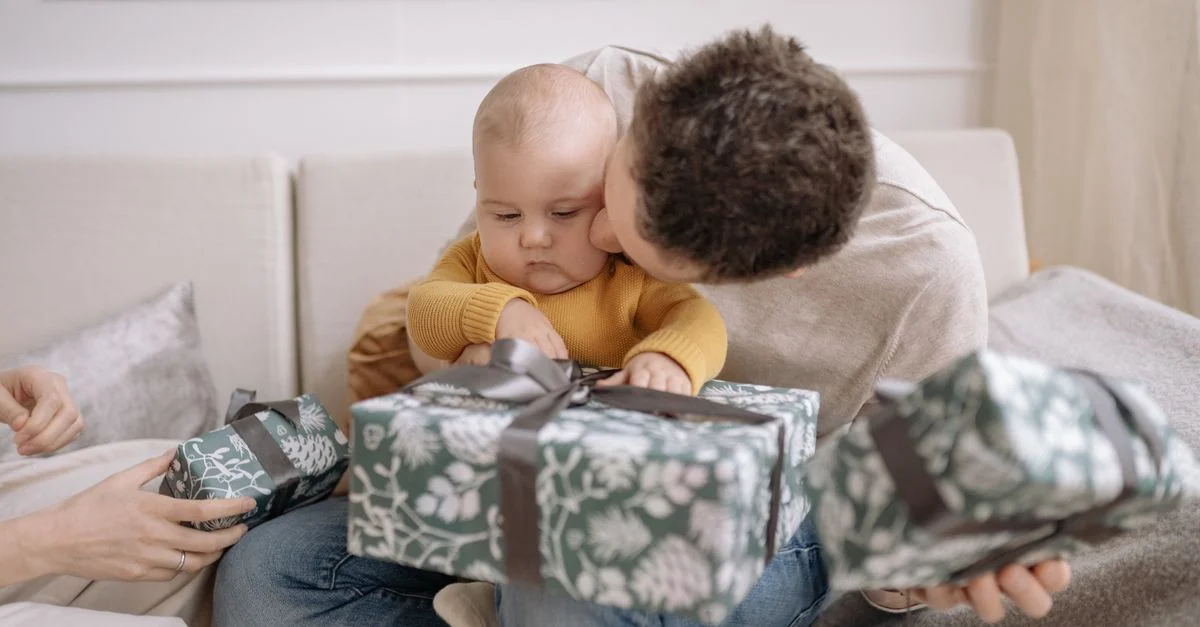 Padre besa a su bebé en el sofá con regalos navideños en el interior, momento familiar cálido.