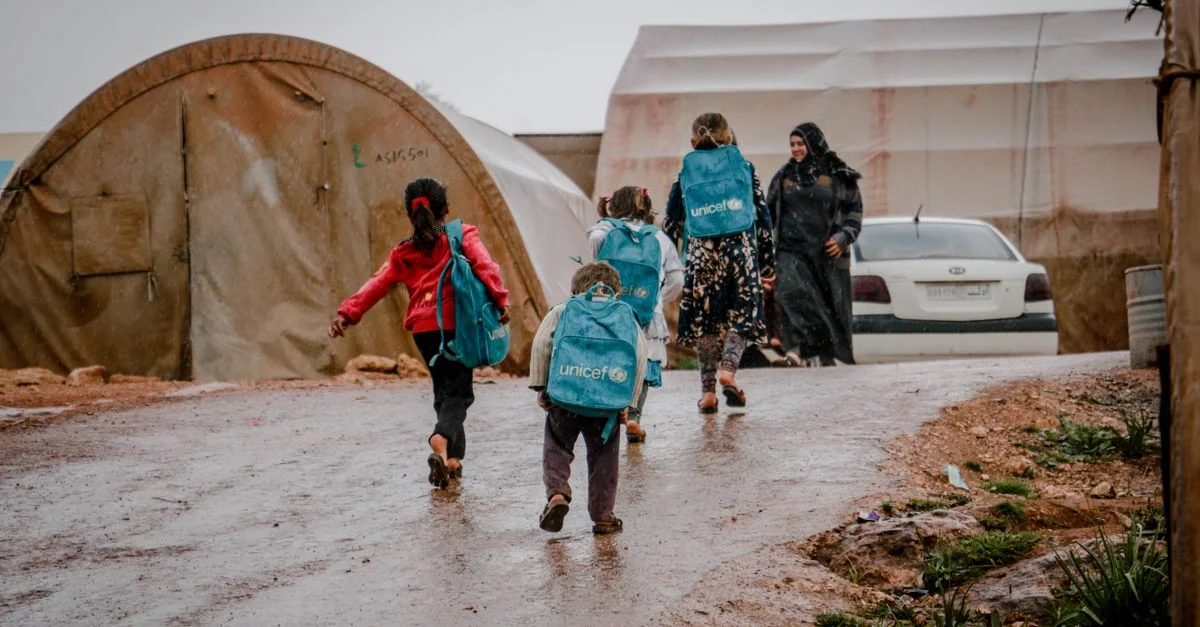Des enfants avec des sacs à dos marchent dans un camp syrien sous la pluie.