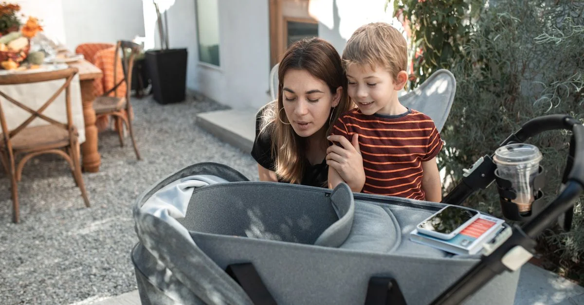 Una madre y su hijo sonriendo y mirando a un bebé en un carrito al aire libre en un día soleado.
