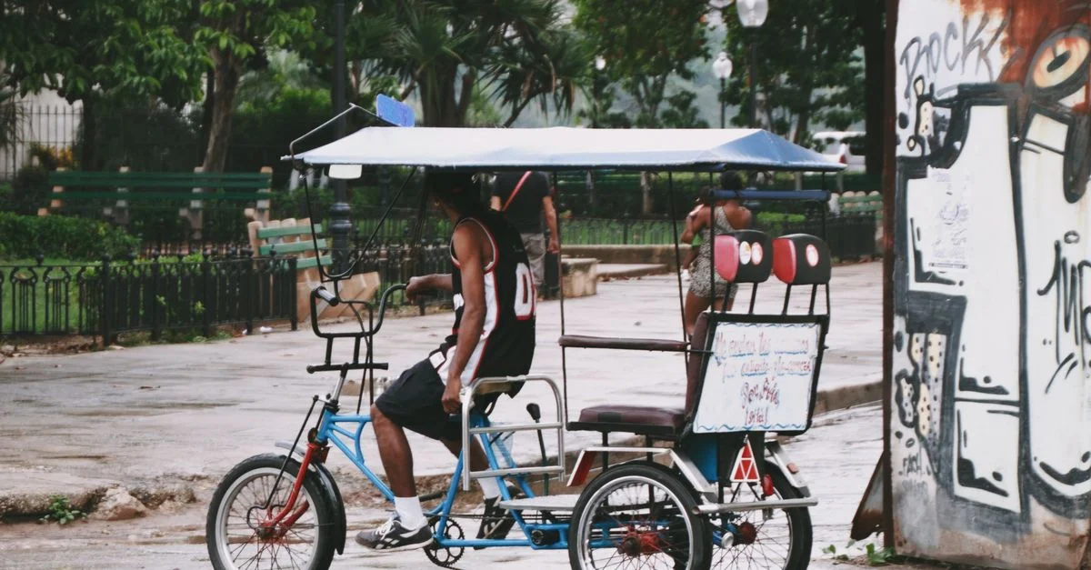 Un hombre se sienta en un rickshaw en una escena de parque urbano con graffiti y vegetación exuberante.