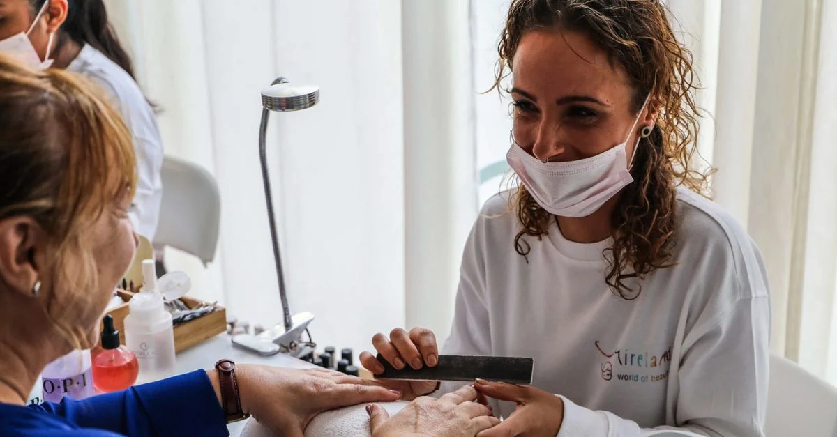 Una esteticista con mascarilla manicura las uñas de una clienta en un salón luminoso.