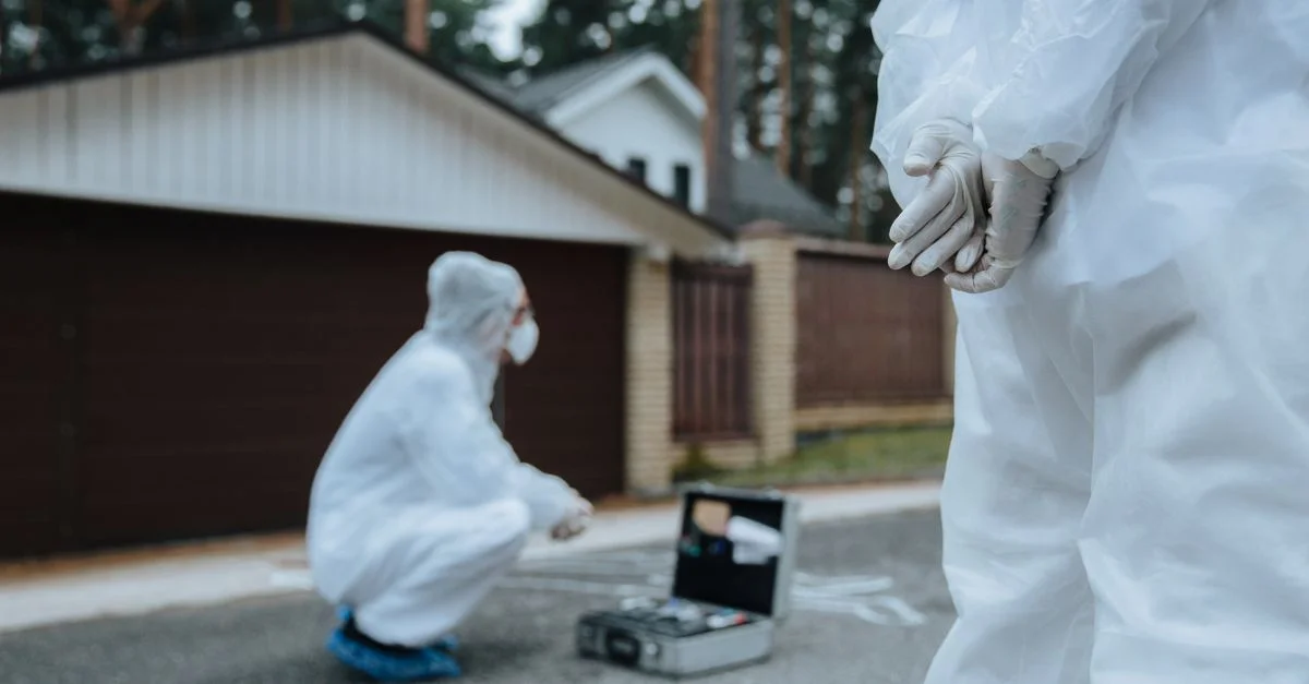 Des enquêteurs légistes en costumes blancs et masques examinant une scène de crime en dehors d'un quartier résidentiel.
