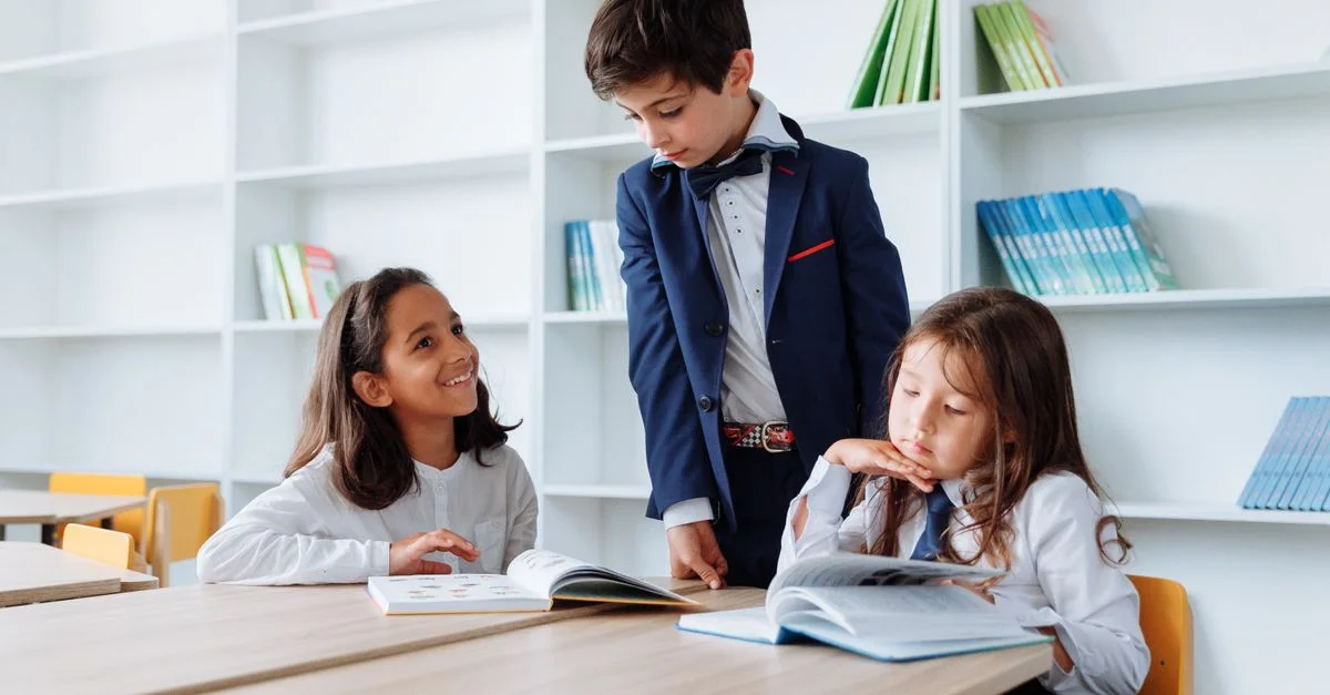 Tres niños participando en actividades de lectura y estudio en una mesa de la biblioteca con estanterías de libros.