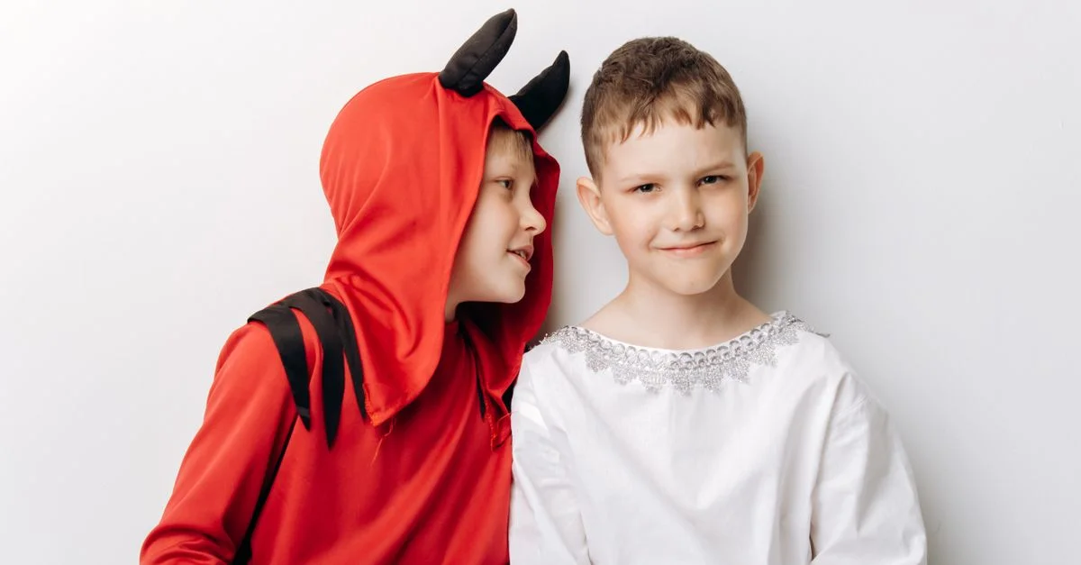 Dos niños disfrazados de un ángel y un diablo para Halloween posan humorísticamente frente a un fondo simple.