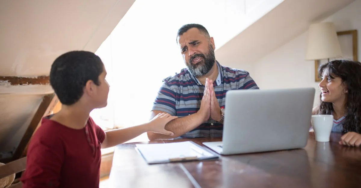 Un padre interactúa con sus hijos en casa, compartiendo sonrisas y conversaciones alrededor de un portátil.