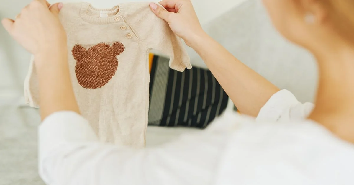 Femme examinant un pull beige pour bébé avec un design d'ours en peluche, mettant en valeur des vêtements pour bébé mignons.